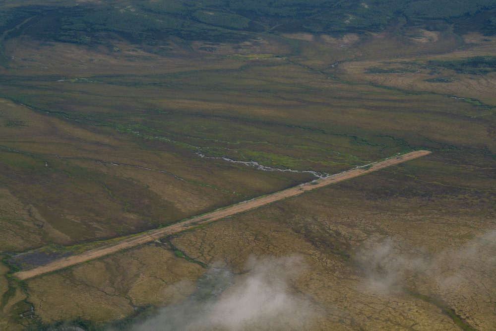 Aerial of a gravel airstrip laid in a straight line across open tundra, with cloud below the plane