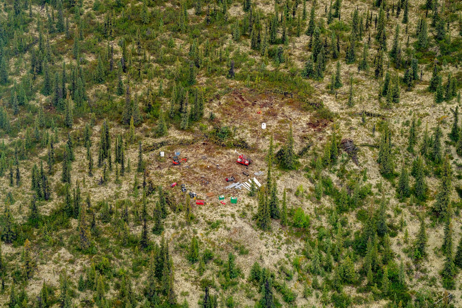 Aerial looking down on a cleared industrial pad with equipment and fuel drums cut into open spruce forest