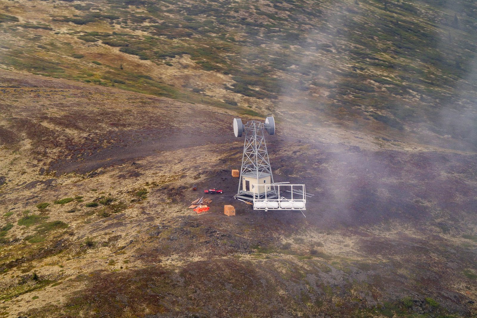 A communications tower with microwave dishes, a small building and equipment on open tundra, seen from the air