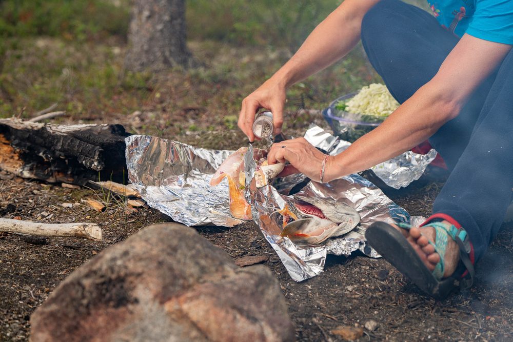 Hands opening foil-wrapped fish on the coals of a shore fire