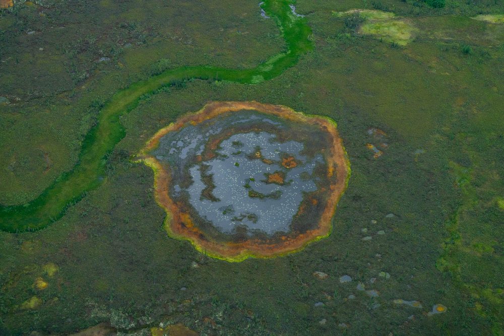 Aerial of a tundra pond ringed in rust orange where iron stains the waterline