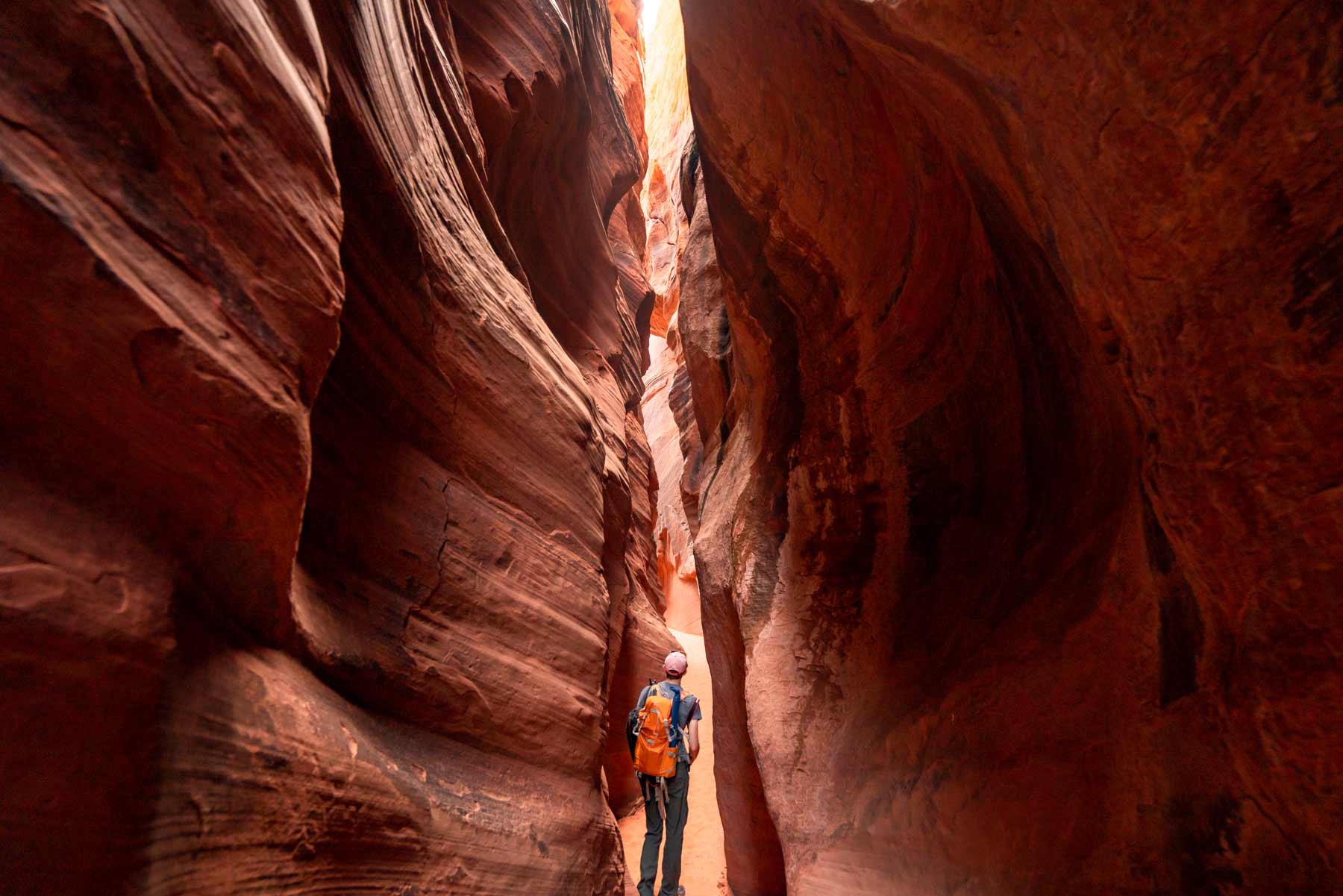 A hiker with an orange pack looking up a narrow red-rock slot canyon