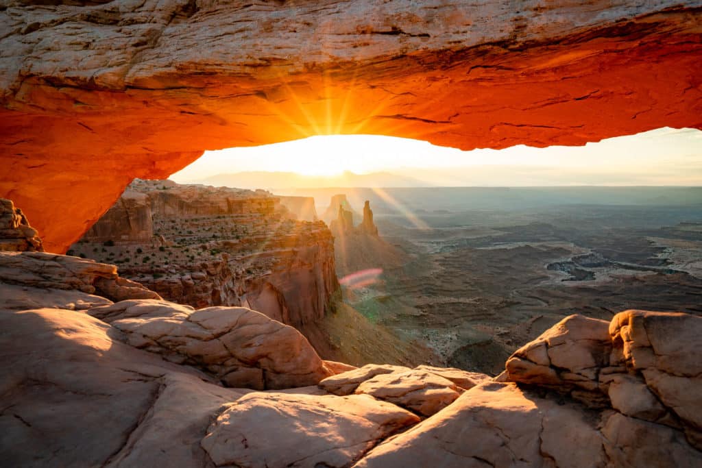 Sun peeking over the tops of mountains as seen through mesa arch in Canyolands National Park, Utah.