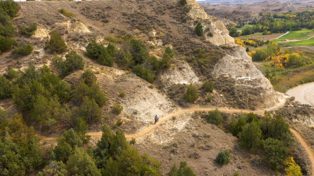 A mountain biker on the Maah Daah Hey Trail above the Little Missouri River, gold cottonwoods along the water below the badlands.
