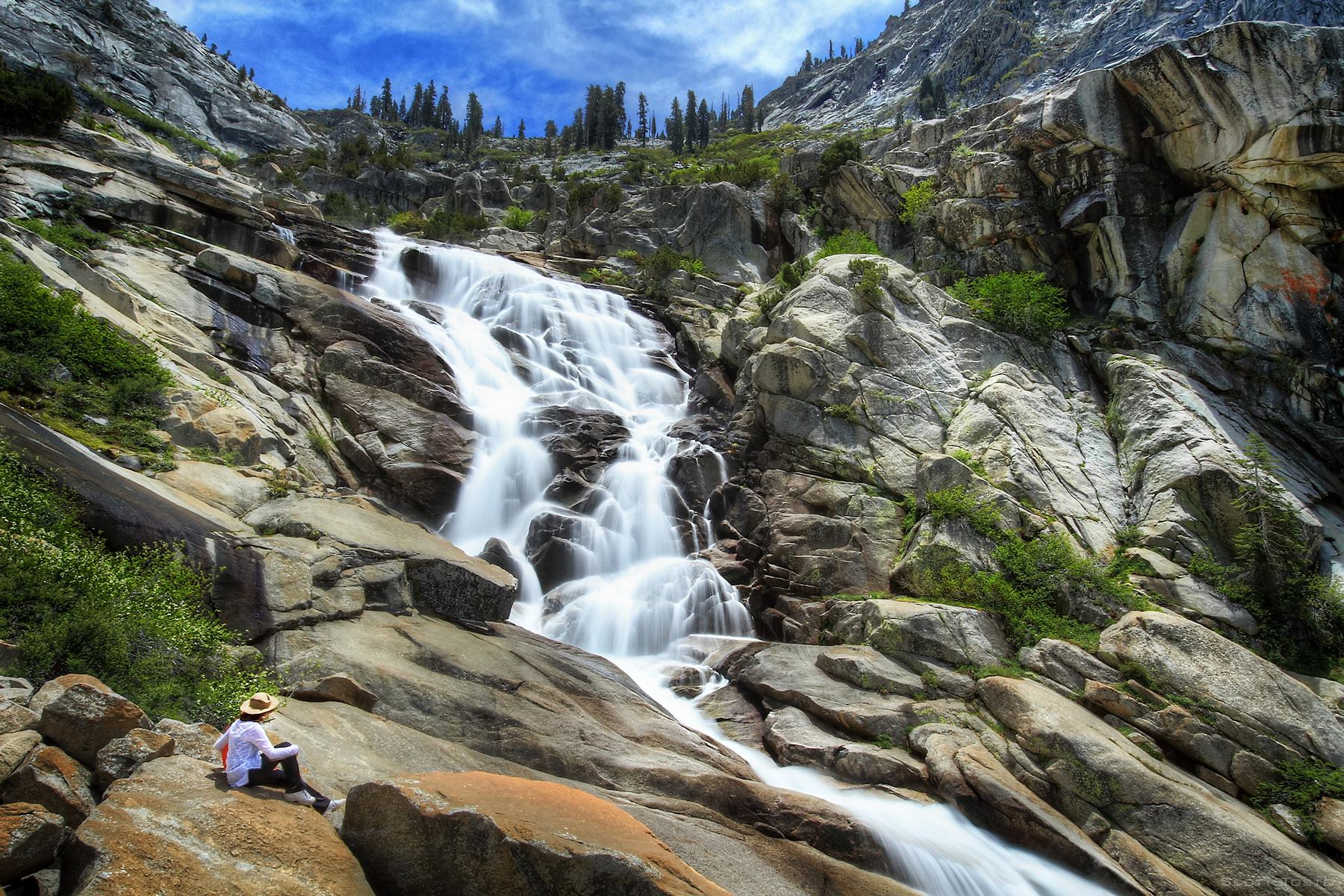 The cascading waterfall of Tokopah Falls comes down a granite stairstep with alpine trees up top and a woman sitting on slickrock below. 