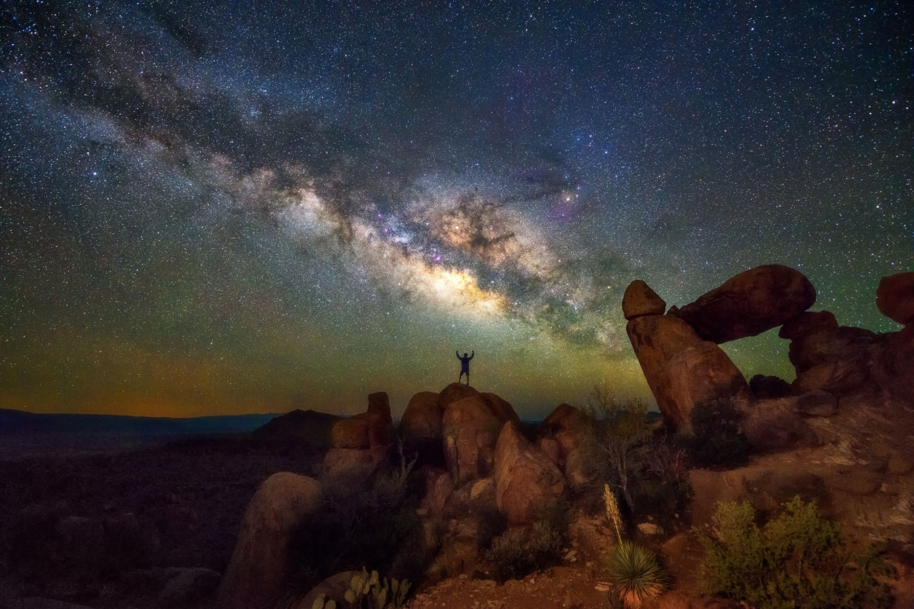 A person stands on a rock with the Milky Way above him in the night skies. 