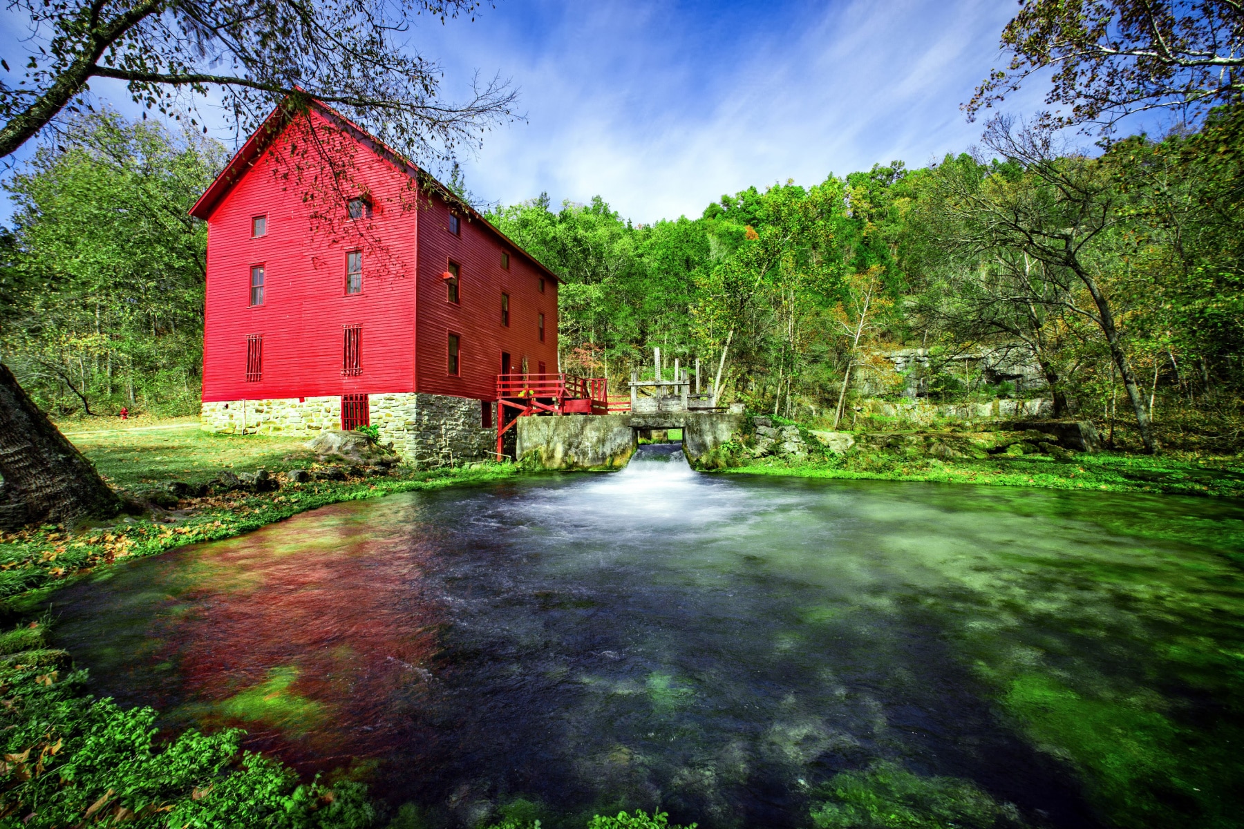 A red mill in Ozark National Scenic Riverways sits along the west side of the river surrounded by trees. 
