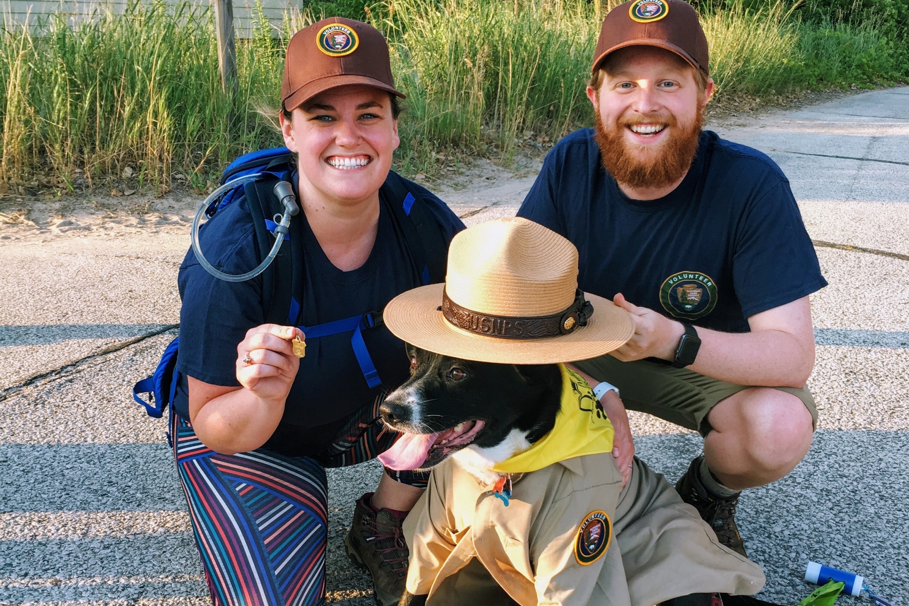 Two NPS volunteers with a dog wearing a ranger hat. 
