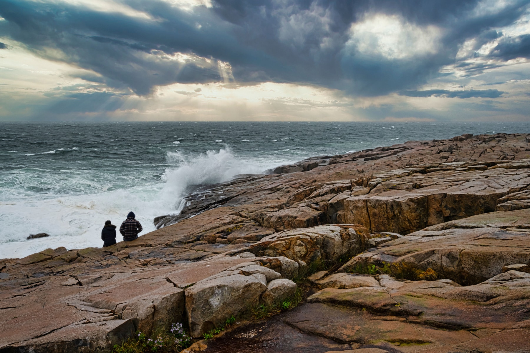Two people sit on a rocky outcropping watching waves crash under stormy skies.