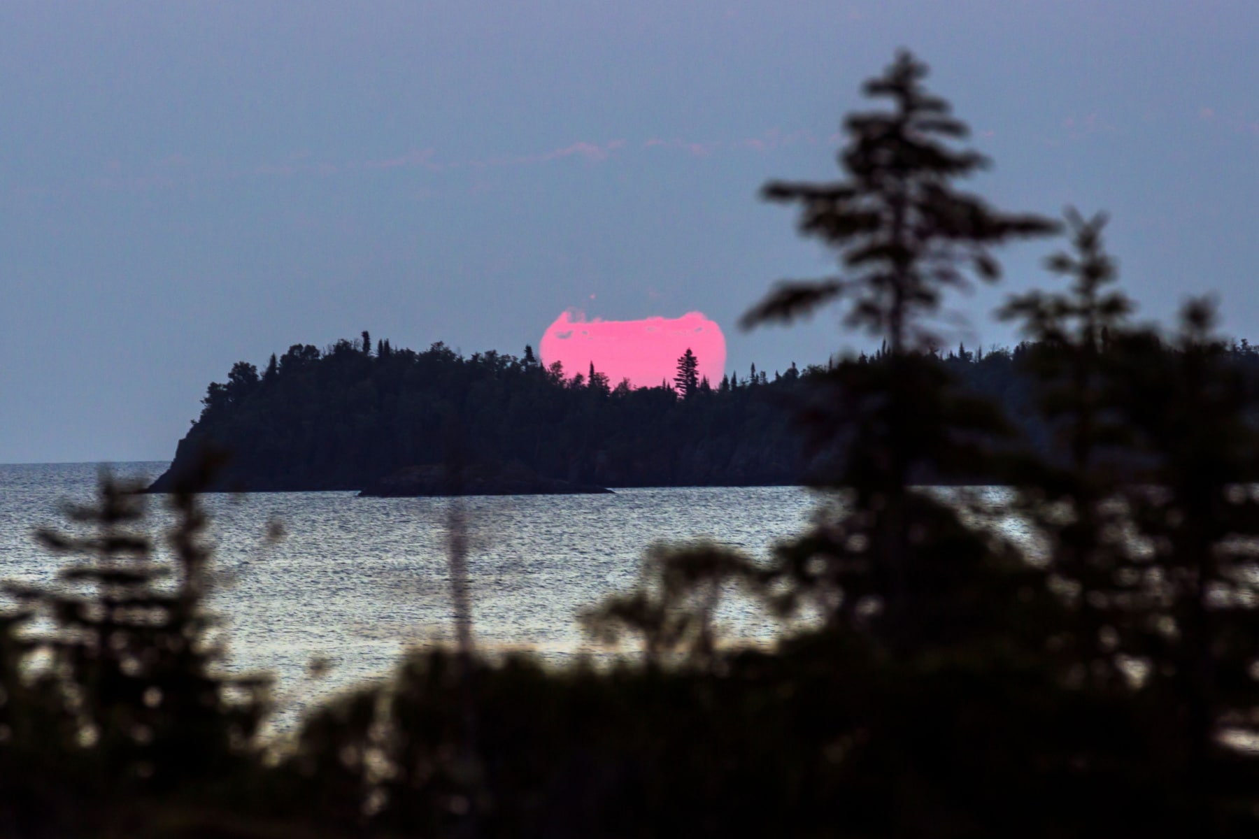 The sun sets with a pink hue on a dark blue sky behind an island in Lake Superior Michigan.