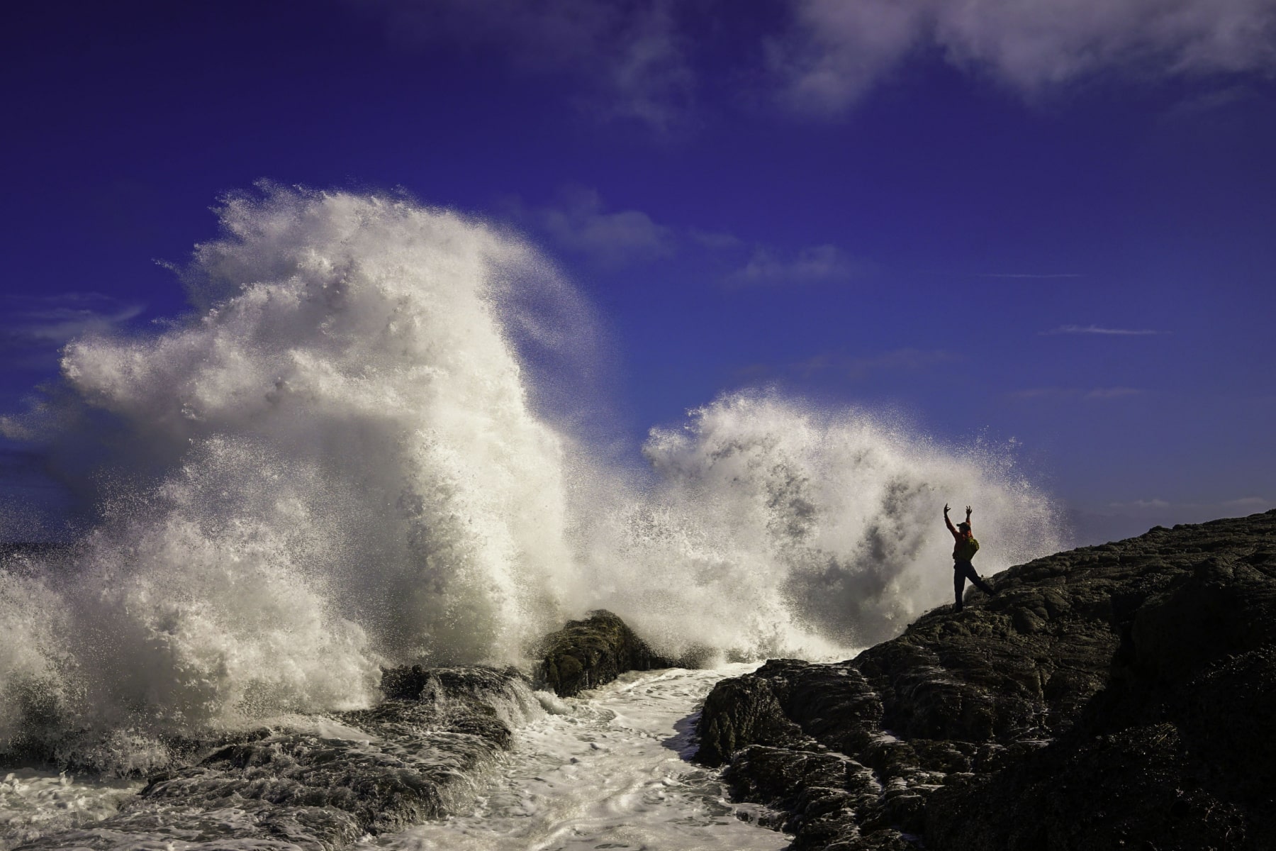 A huge wave crashes on rocks while a surfer on the rocks raises him arms at Channel Islands National Park. 