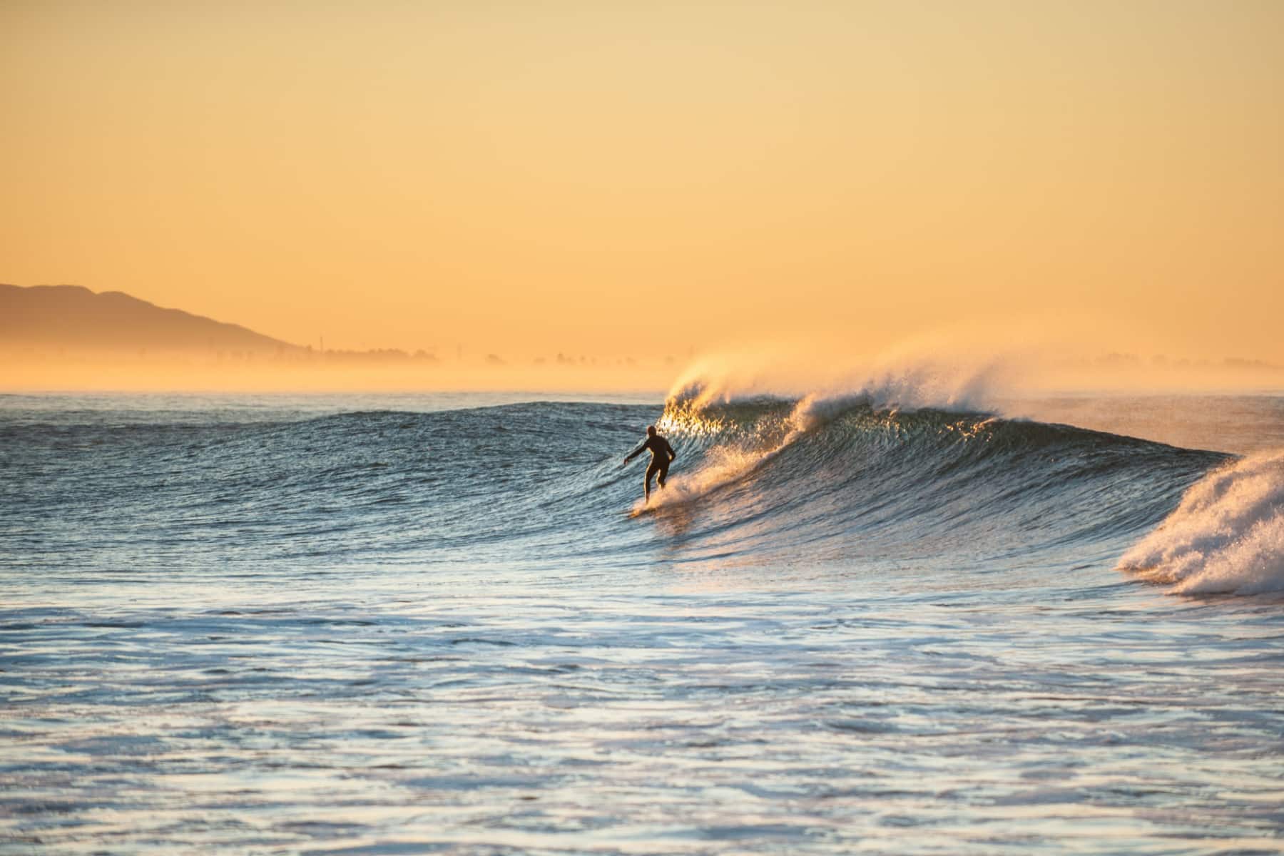 A surfer riding a small wave at Ventura Beach in the early morning glow.
