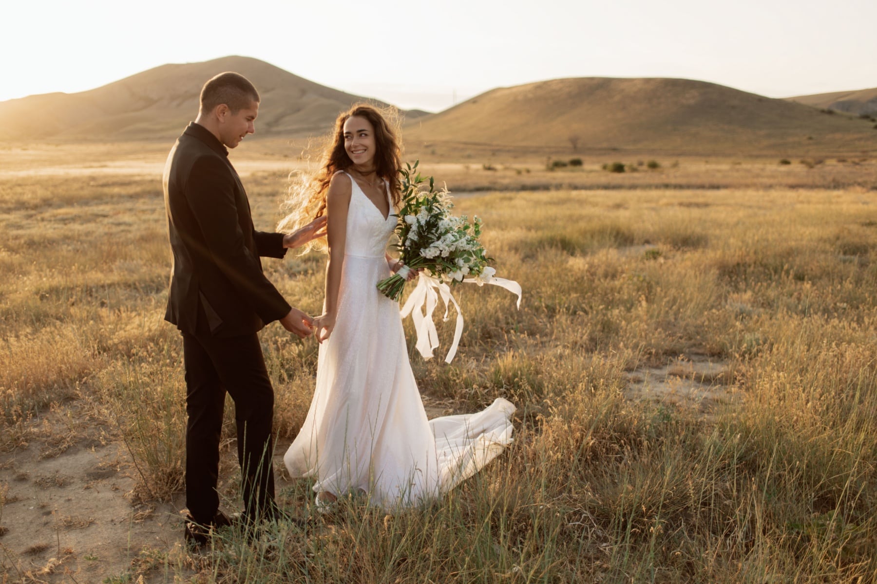 A bride and groom stand in a grassy field with hills behind them.