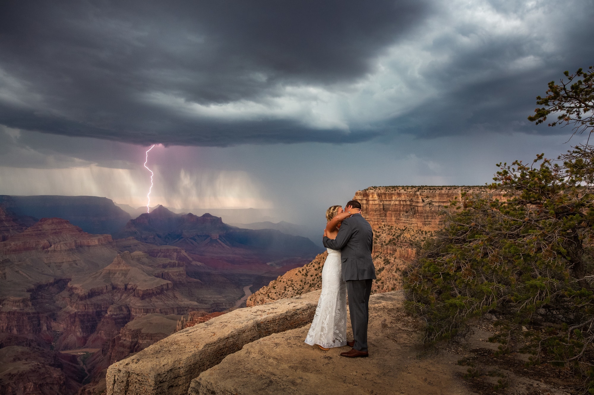 A couple embrace at the edge of the Grand Canyon with lightning in the background.