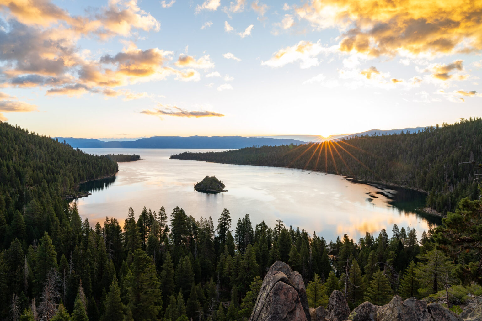 Emerald Bay and Fannette Island on Lake Tahoe at sunrise, the sun breaking over a ridge of dark conifers.